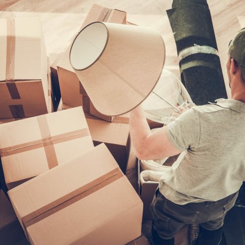 Caucasian Divorced Men in His 30s Moving Out From His Home. Staying Between Cardboard Boxes Preparing to Pack His Lamp.